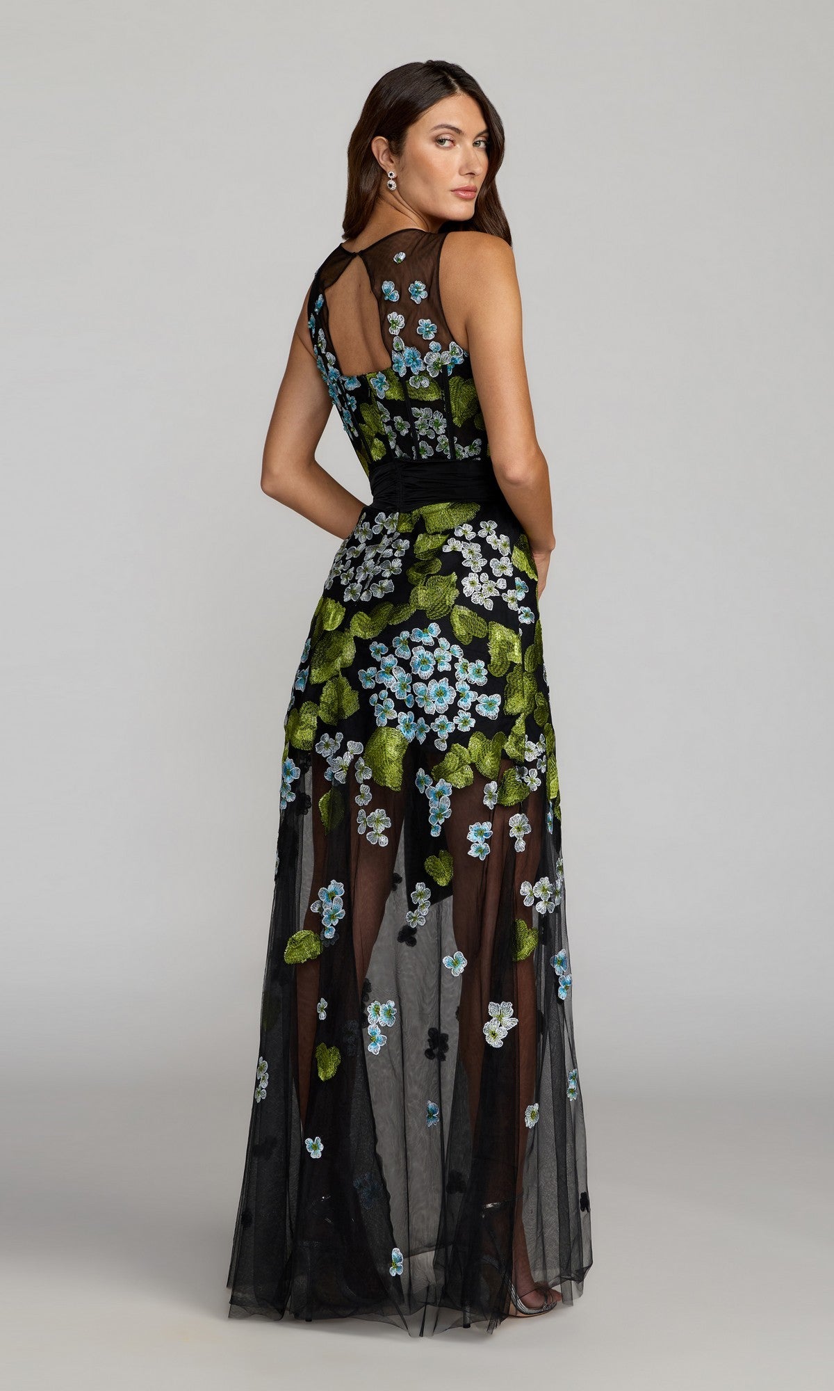 A woman wears the Mac Duggal 21058 long formal dress featuring sleeveless styling, blue and green floral embroidery, and a statement waist bow, standing and looking over her shoulder.